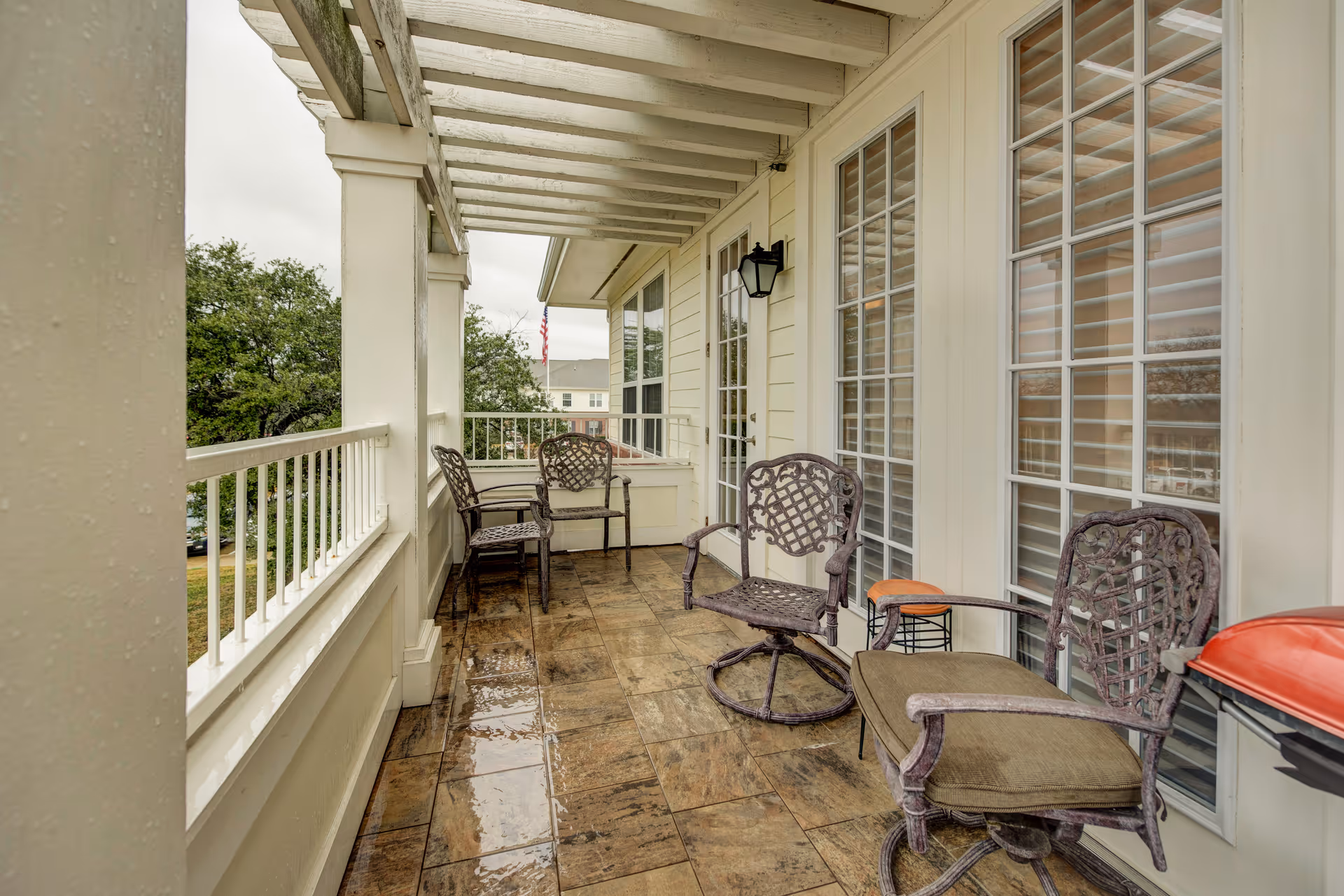 Covered balcony with several metal chairs, a small table and wet tiled floor overlooking trees and nearby buildings.
