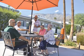 Five elderly people sitting and standing around a round outdoor table with an umbrella, enjoying a sunny day at Stonewall Gardens Assisted Living with mountains and palm trees in the background.
