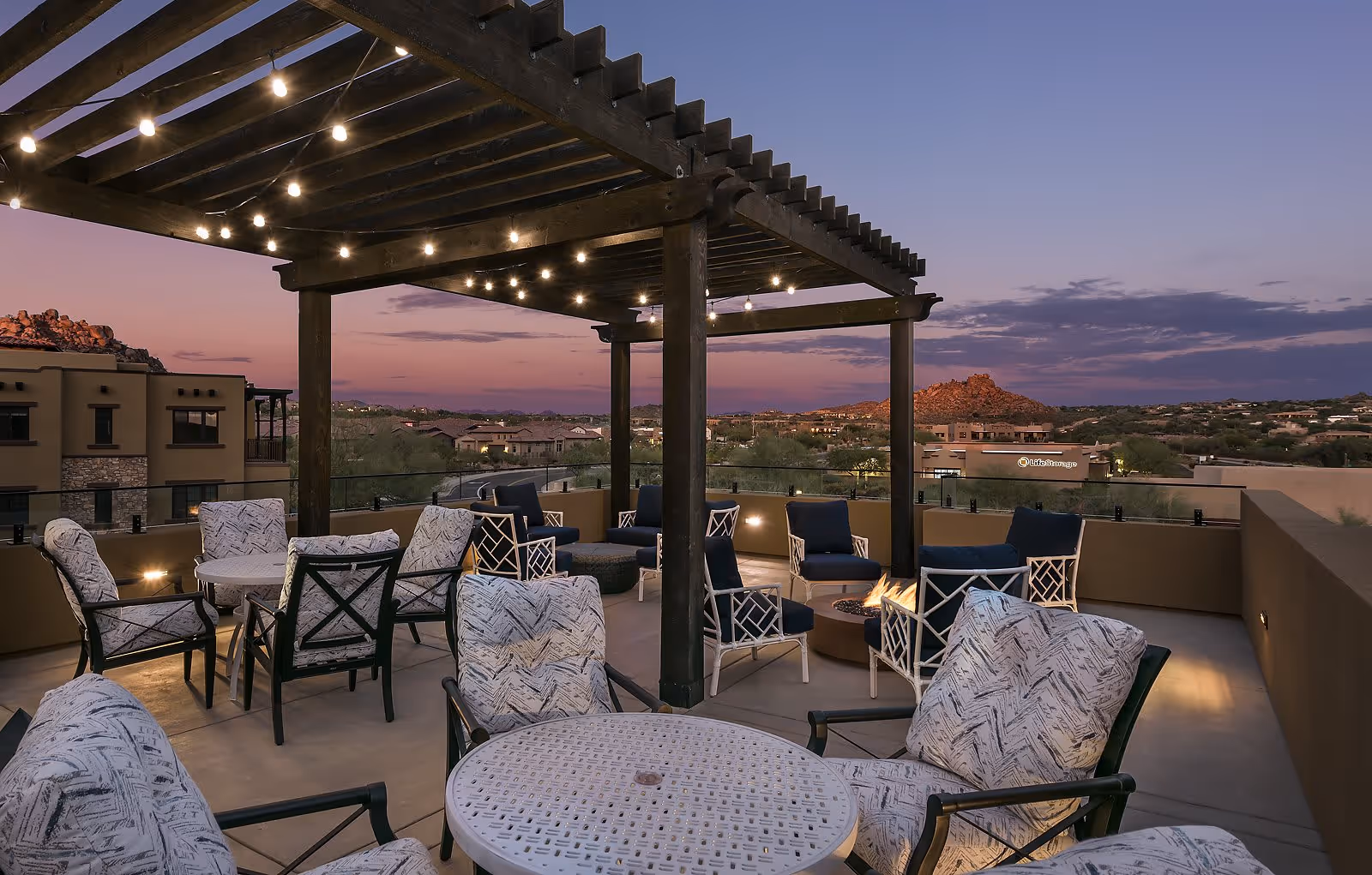 Outdoor patio area at dusk with cushioned chairs and tables under a pergola adorned with string lights, overlooking a scenic view of distant hills and buildings.