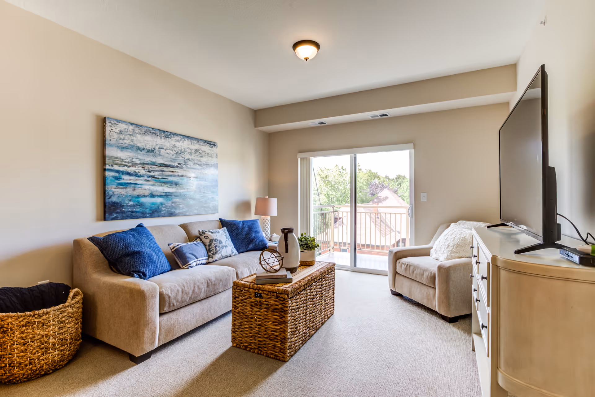 A cozy living room with a beige sofa adorned with blue and patterned pillows, a wicker coffee table with decorative items, a beige armchair, a large flat-screen TV on a light-colored dresser, and a sliding glass door leading to a balcony with a view of trees and rooftops.
