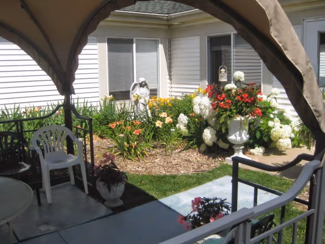 View from a shaded gazebo overlooking a courtyard garden with potted flowers, a white plastic chair, an urn of red and white blooms, a small statue, and the building's exterior windows.