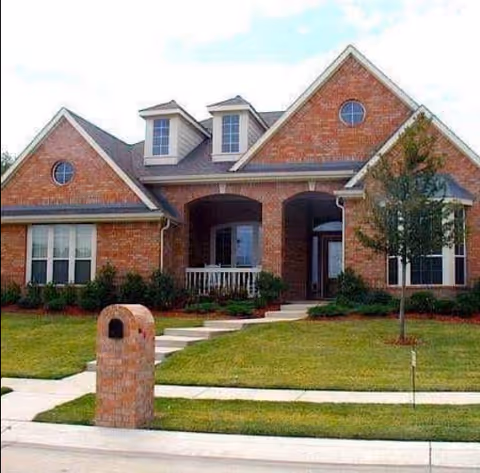 A single-story brick house with a front porch, two dormer windows on the roof, a circular window on the front gable, a small tree on the right side, and a brick mailbox at the edge of the sidewalk. The lawn is neatly maintained with a concrete walkway leading to the front door.