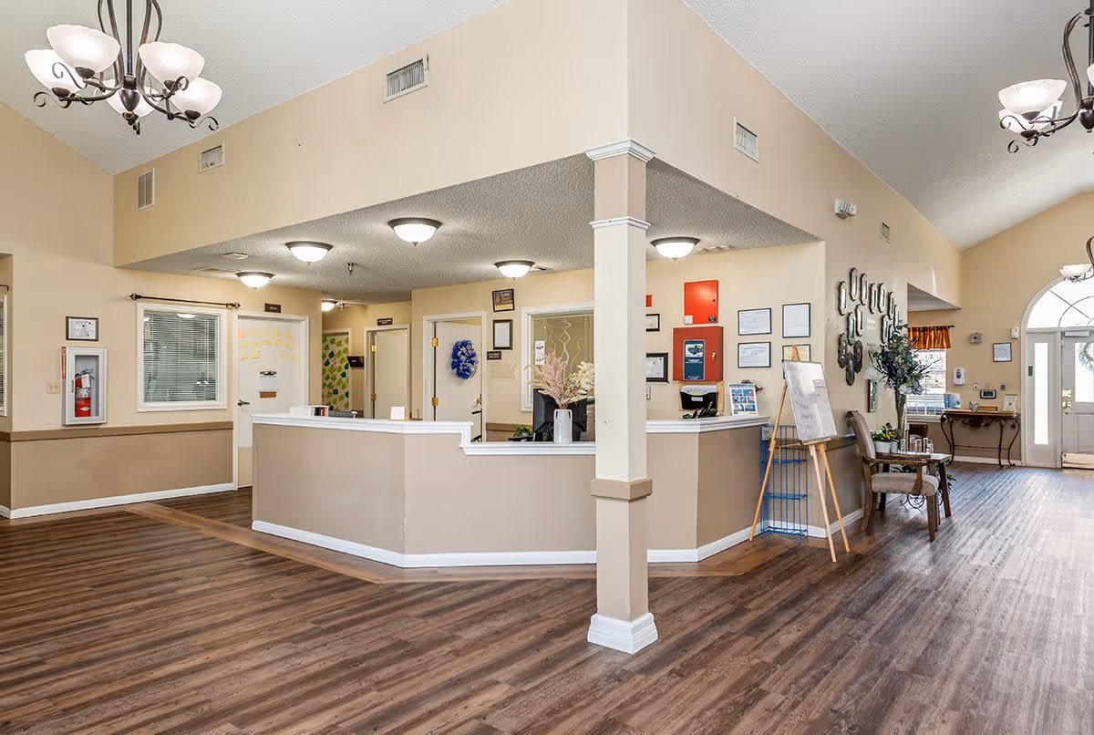 Reception area inside Lucas Court facility with a beige and white front desk, wood flooring, beige walls, ceiling lights, and decorative elements including flowers, framed certificates, and a seating area with chairs near the entrance door.
