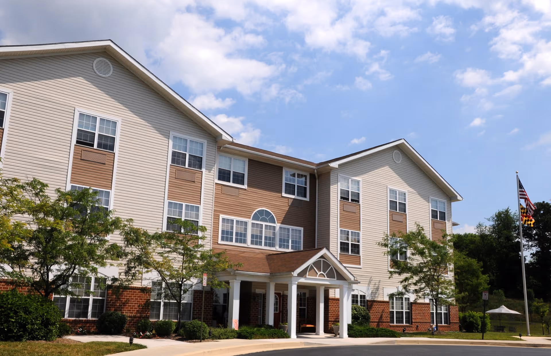 Front exterior of a multi-story senior living building with a covered entrance, landscaping, and a flagpole under a blue sky.