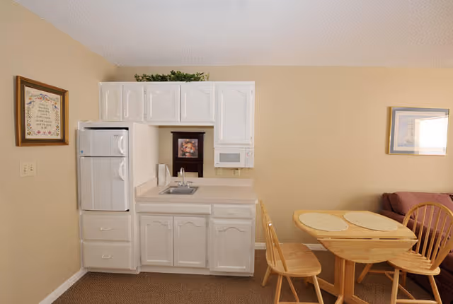 Small kitchen area with white cabinets, a refrigerator, a microwave, and a sink. Adjacent to the kitchen is a wooden dining table with four chairs and placemats. The walls are beige, and there are framed pictures hanging on the walls.