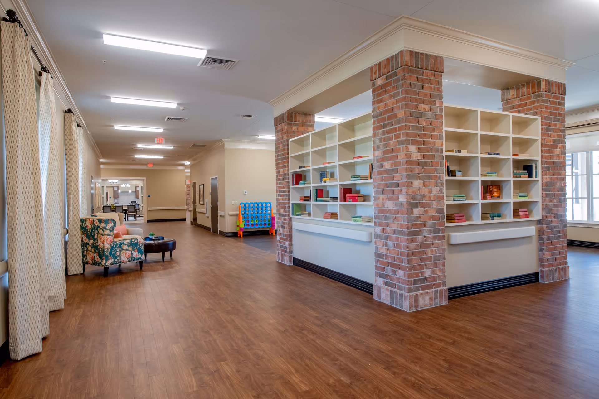 A spacious interior hallway in an assisted living facility with wood flooring, brick pillars surrounding a white bookshelf filled with books, floral armchairs, and large windows with curtains allowing natural light.