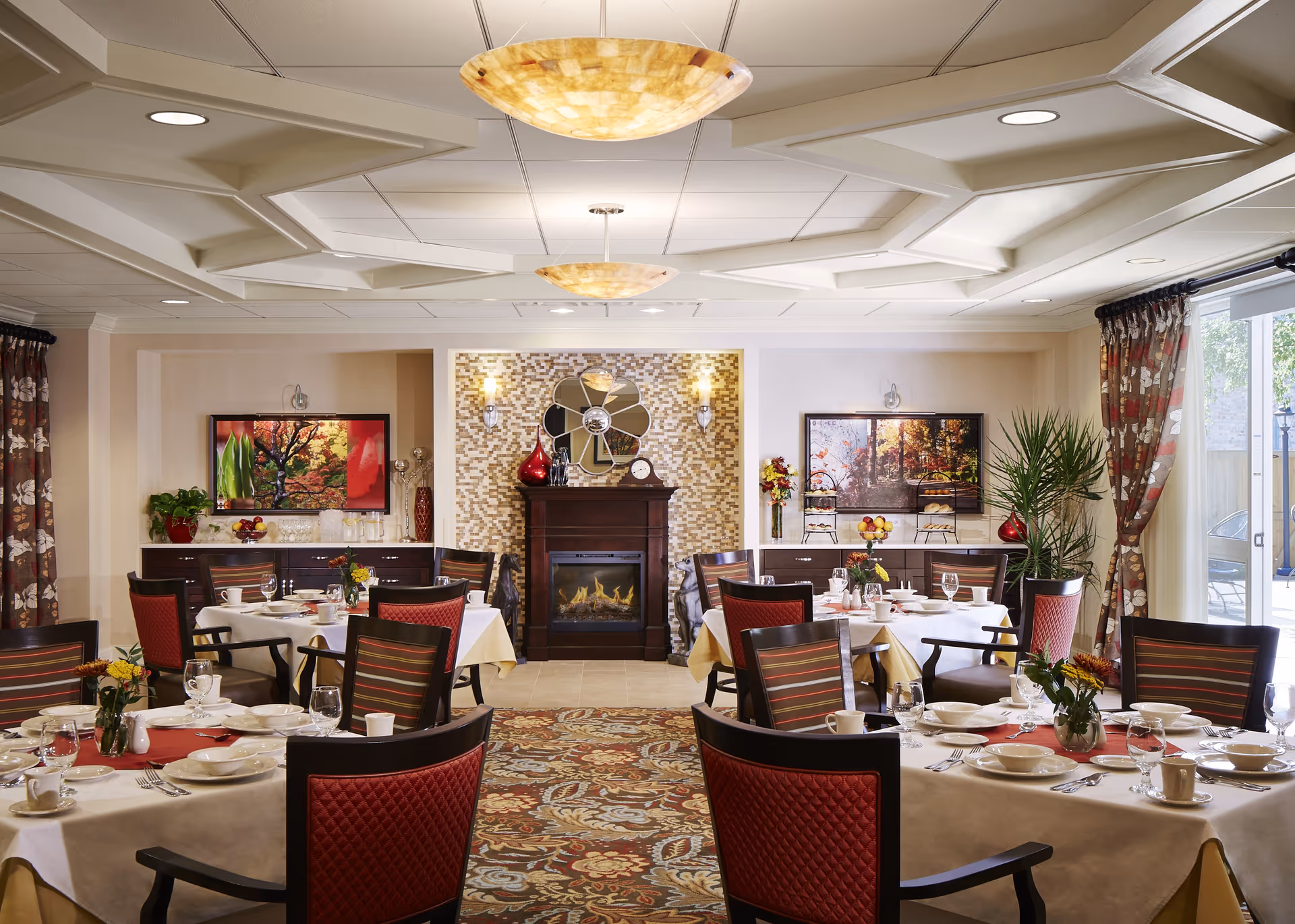 A well-lit dining room with several round tables covered in white tablecloths and set with plates, cups, glasses, and silverware. The room features a decorative fireplace with a mirror above it, two framed pictures on the walls, floral curtains, and a patterned carpet. There are red and brown cushioned chairs around the tables, and plants and decorative items are placed on sideboards along the walls.