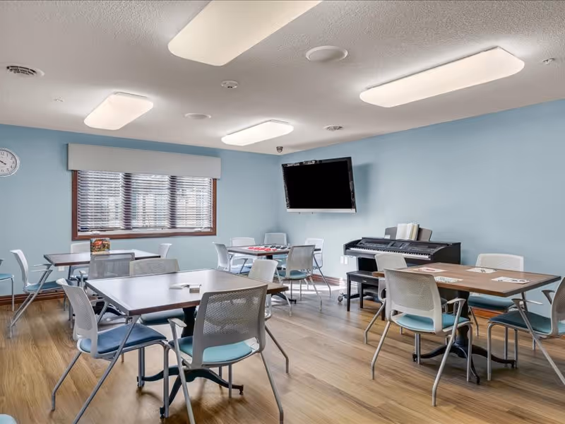 A bright room with light blue walls and wooden flooring featuring multiple tables and chairs arranged for group activities. A digital piano is placed against one wall with sheet music on it, and a large flat-screen TV is mounted on the wall above the piano. A window with blinds allows natural light into the room, and ceiling lights provide additional illumination.