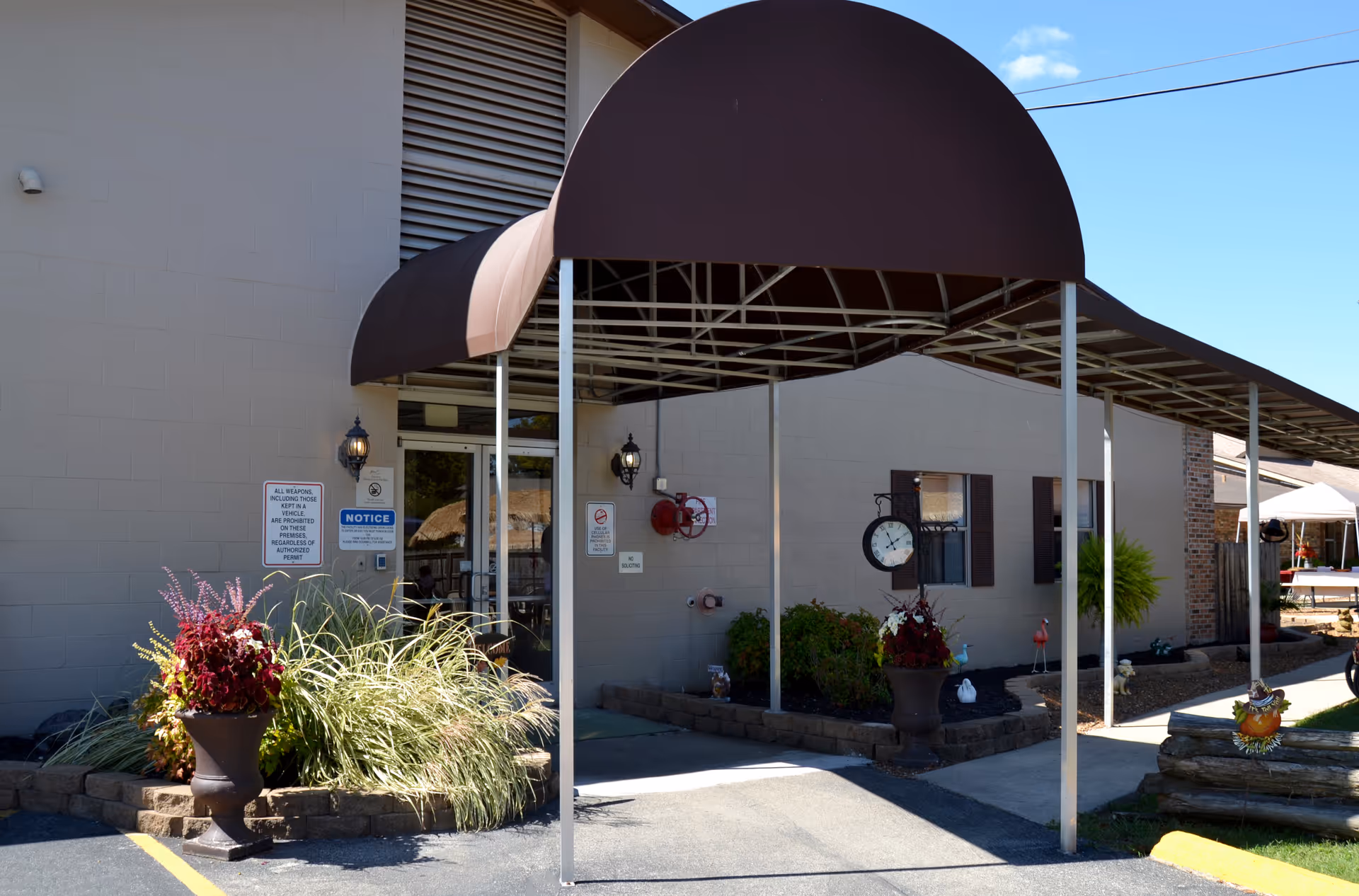 Exterior entrance of a care facility with a maroon covered canopy, potted plants, and signage on the building.