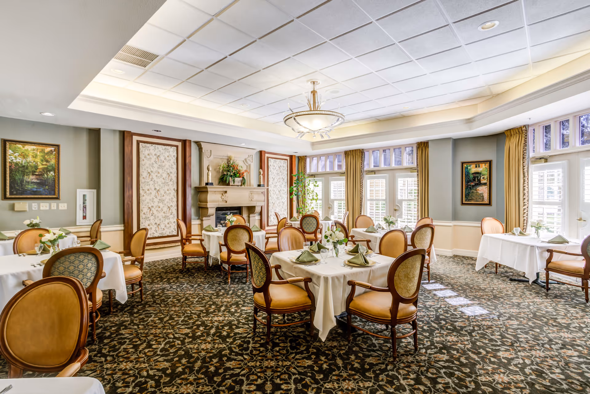 A bright and elegant dining room with round and square tables covered in white tablecloths, set with green napkins and glassware. The room features upholstered wooden chairs, a decorative fireplace, large windows with yellow curtains, and framed artwork on the walls. The carpet has a detailed floral pattern.