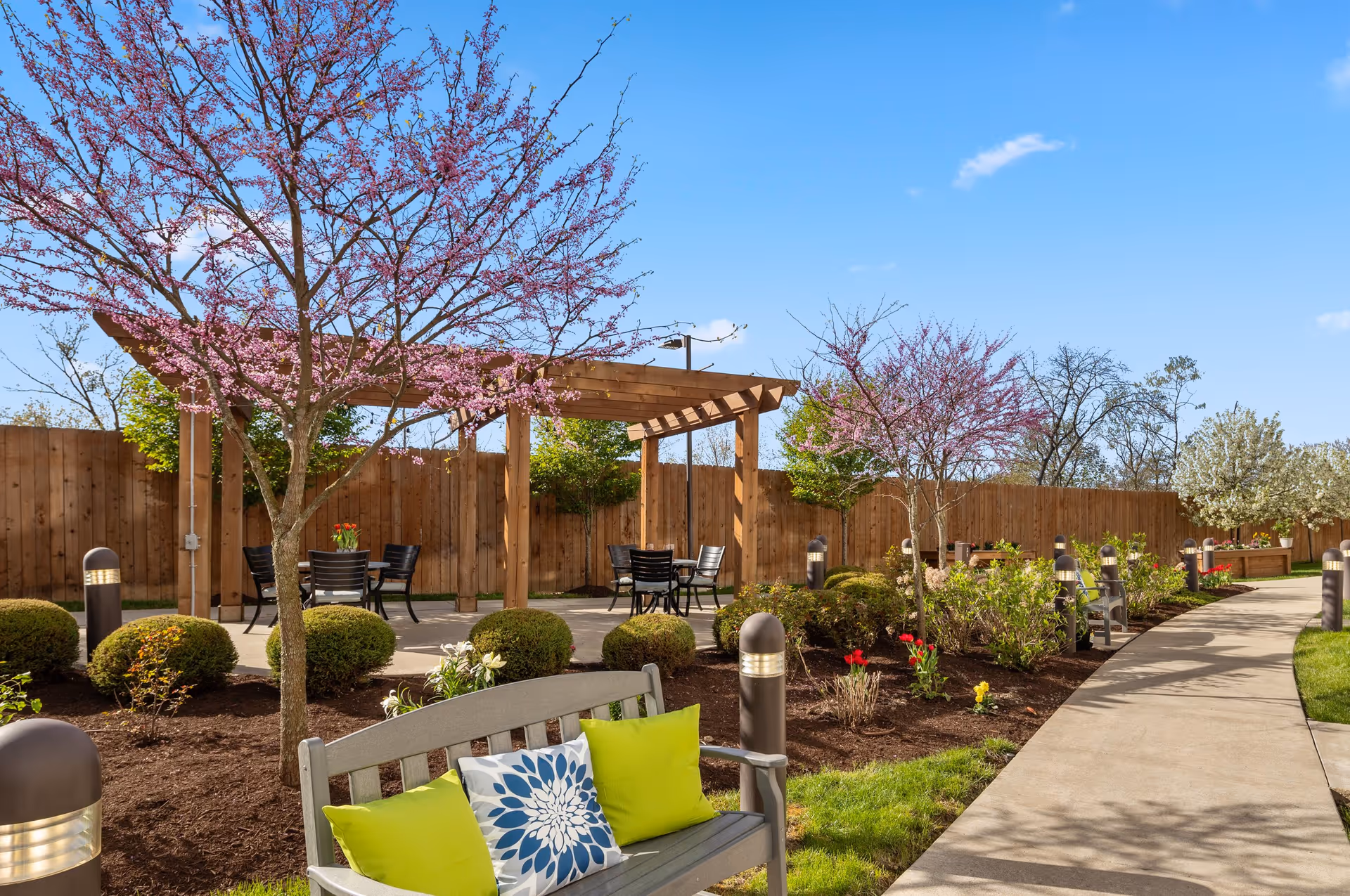 A peaceful outdoor garden area at The Remington of McCandless featuring a wooden pergola with black chairs and tables underneath. There are blooming pink trees, neatly trimmed bushes, and colorful flowers along a curved concrete pathway. A wooden bench with green and blue floral cushions is in the foreground, and the area is enclosed by a wooden fence under a clear blue sky.