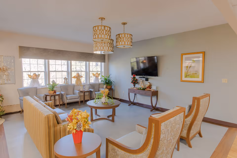 A bright and cozy living room with large windows letting in natural light. The room features a striped sofa, two patterned armchairs, and three gray armchairs arranged around wooden coffee and side tables. Decorative plants and floral arrangements are placed on the tables. A flat-screen TV is mounted on the wall above a console table with holiday decorations. Two pendant lights hang from the ceiling, and a framed picture is on the wall.