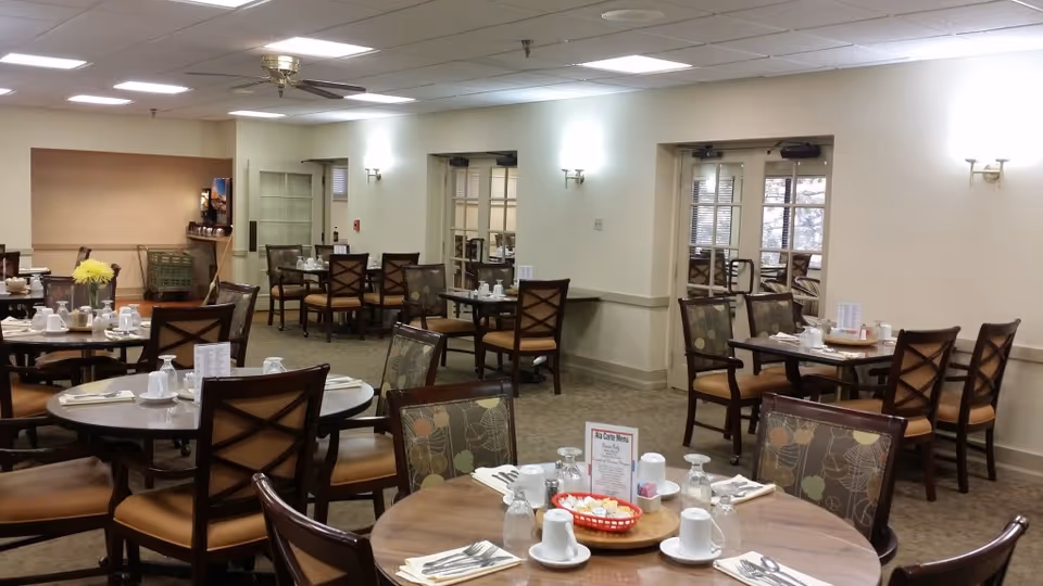 A dining room with multiple round and square tables set with white cups, glasses, napkins, and silverware. Chairs with patterned and plain cushions surround the tables. The room has beige walls, carpeted floor, ceiling lights, and a ceiling fan. There are double doors with glass panes on the right side and a beverage station in the back corner.