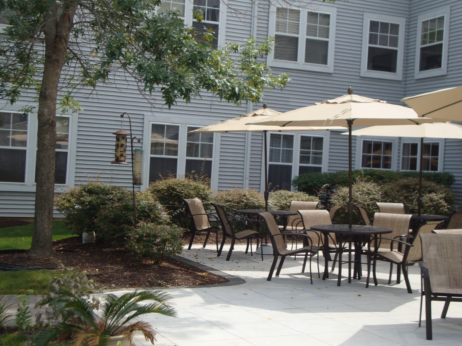 Outdoor patio area with several round tables and chairs under large beige umbrellas, surrounded by bushes and trees, adjacent to a building with multiple windows.