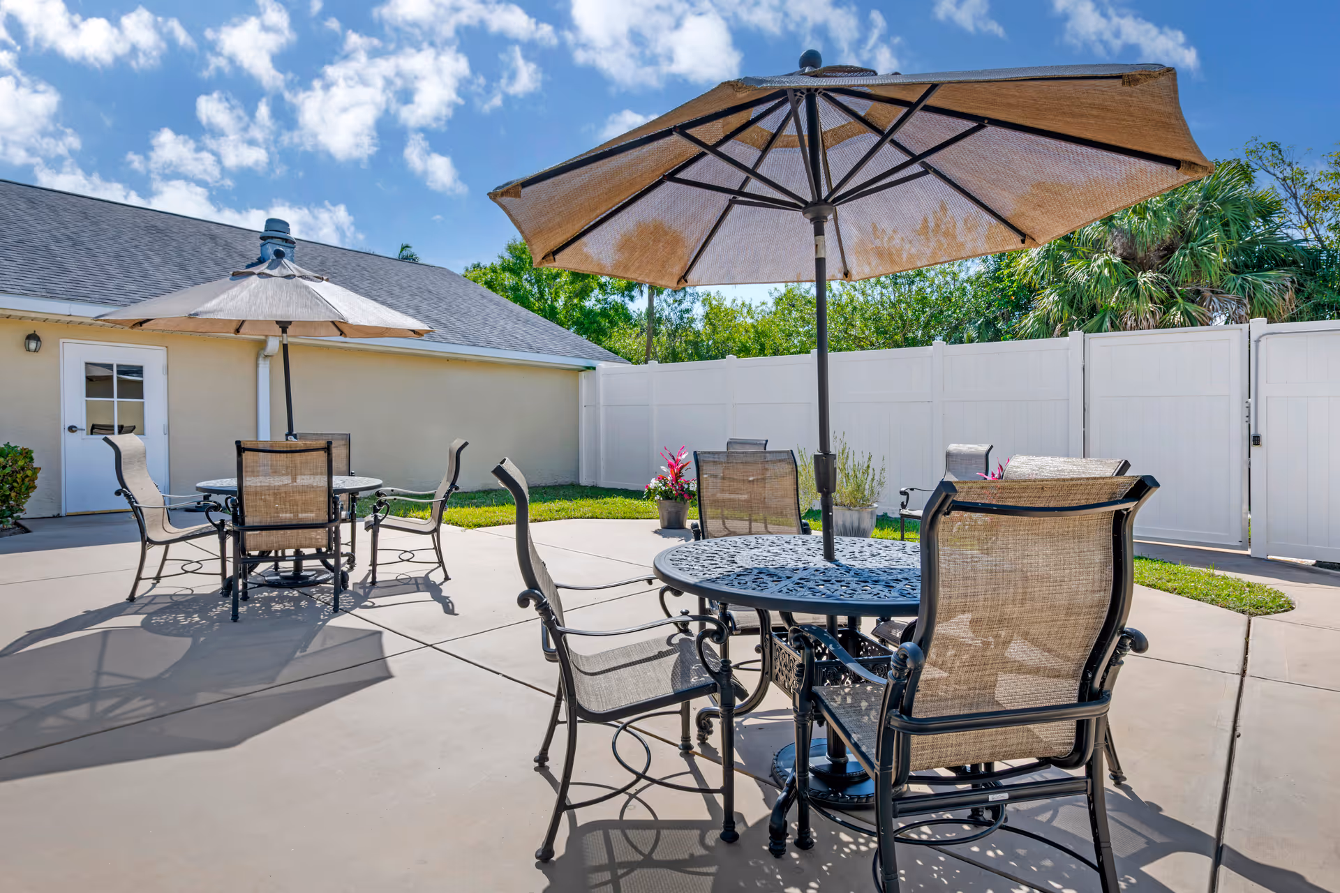 Outdoor patio area with two round metal tables, each surrounded by four mesh chairs. Each table has a large beige umbrella providing shade. The patio is paved with concrete and enclosed by a white fence. There are some potted plants and greenery visible along the fence and a building with a door in the background under a partly cloudy blue sky.