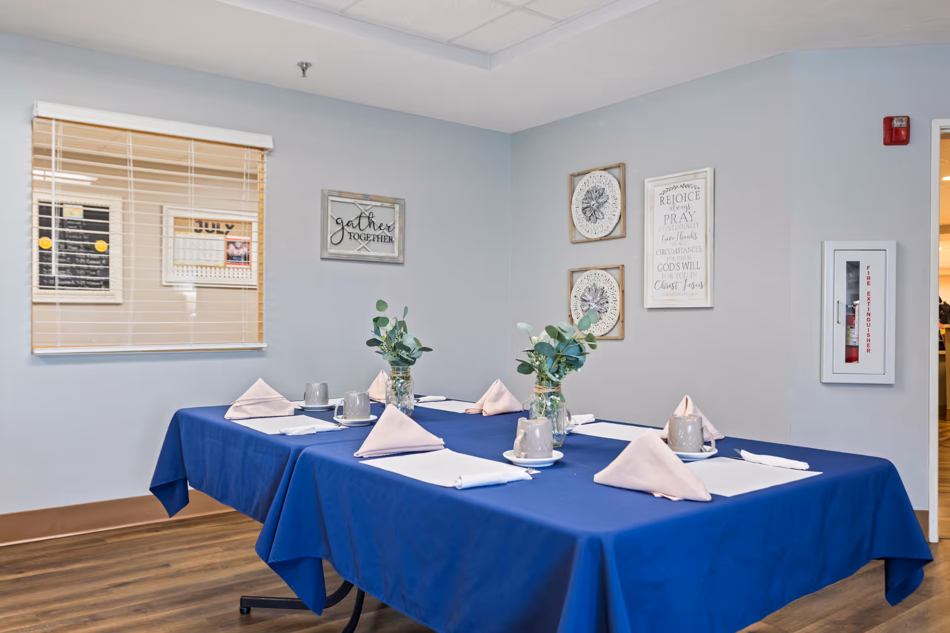 A dining area with a rectangular table covered in a blue tablecloth, set with beige napkins, gray mugs, and white placemats. Two small vases with green foliage are placed on the table. The walls are light gray and decorated with framed artwork and a fire extinguisher cabinet. A window with blinds is visible on the left side.