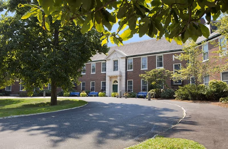 Exterior view of a two-story brick building with a central entrance featuring white columns and a green door, surrounded by trees and greenery with a curved driveway in front.