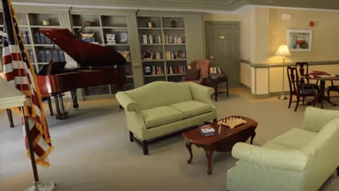 A senior living community common area featuring two light green sofas facing each other with a wooden coffee table in between, a chessboard on the table, a grand piano in the background near a bookshelf filled with books, an American flag on a stand, and a small dining table with chairs and a lamp in the corner.