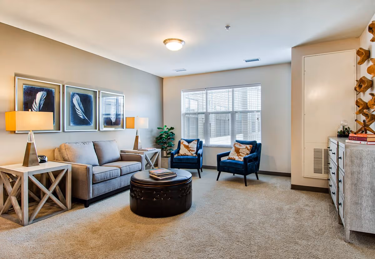 A bright living room with beige walls and carpet, featuring a gray sofa with two side tables and lamps, two blue armchairs with decorative pillows, a round black ottoman coffee table, a large window with blinds, and a dresser with books and decorative items.