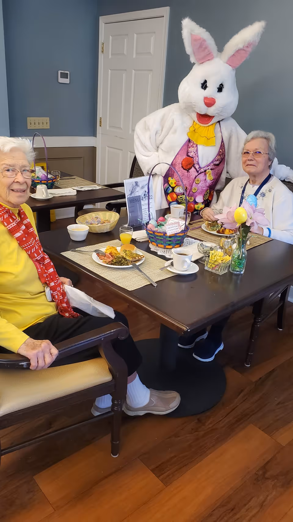 Two seated senior residents eat at a decorated dining table while a person in an Easter bunny costume stands behind them.