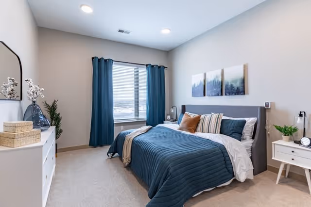 A neatly made bed with a dark blue quilt and multiple pillows in a bedroom with beige walls and carpet. There are three framed pictures above the bed, a window with dark blue curtains, a white dresser with decorative items on the left, and a white nightstand with a plant and lamp on the right.