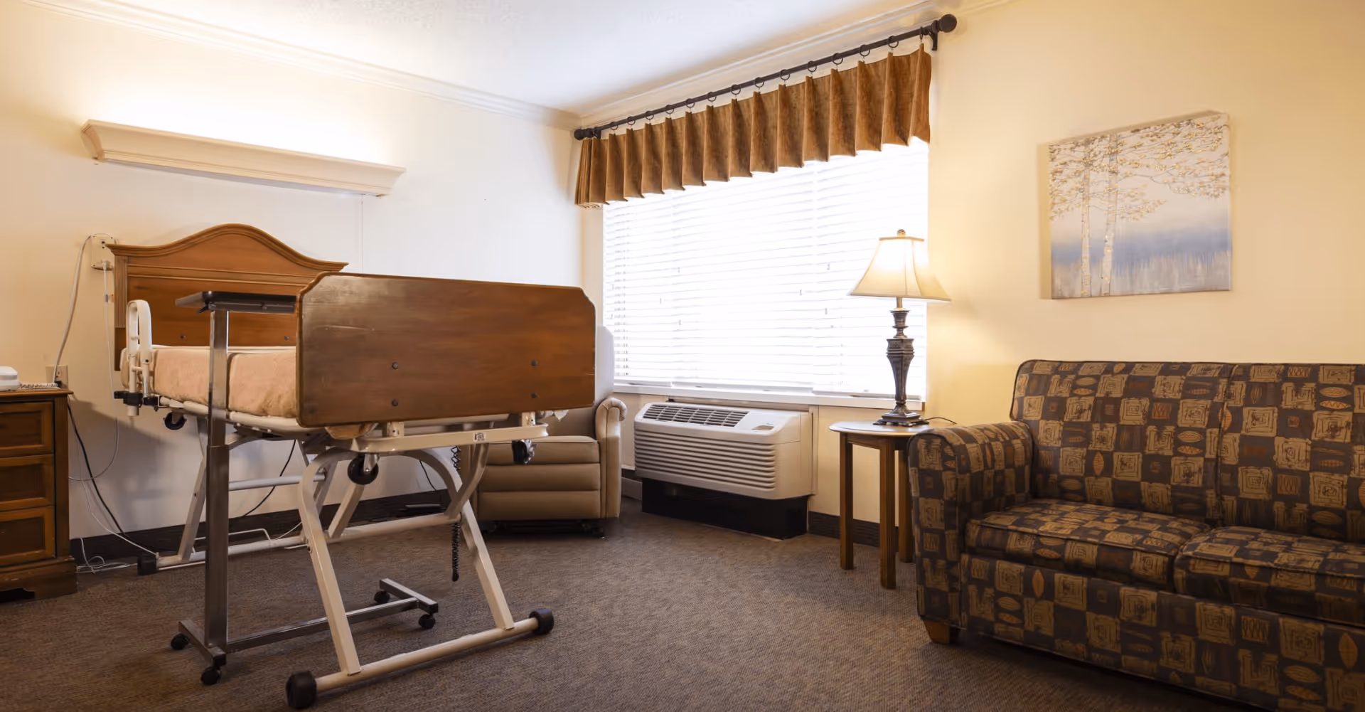 A room in an assisted living facility featuring a hospital-style adjustable bed with wooden headboard and footboard, a patterned sofa, a side table with a lamp, a window with blinds and brown valance, an armchair, and a wall-mounted air conditioning unit.