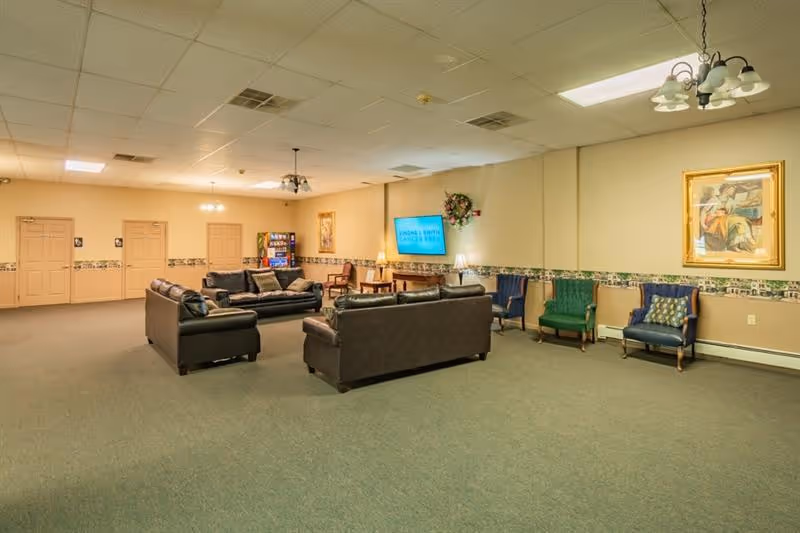 Large common lounge with sofas, armchairs, and a wall-mounted TV in a senior living facility.