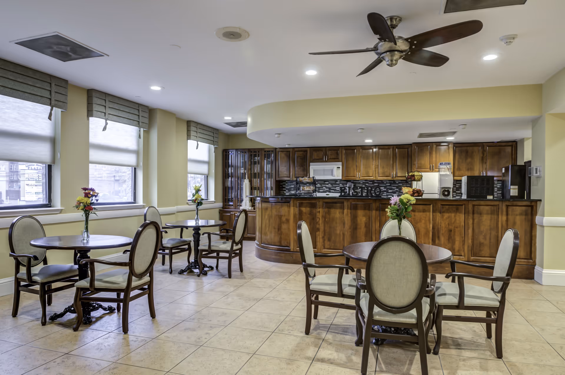 A dining area with round wooden tables and cushioned chairs arranged neatly. Each table has a vase with colorful flowers. The room has large windows with shades, a ceiling fan, and a kitchen area with wooden cabinets, a microwave, and a refrigerator. The backsplash behind the counter has the word 'BISTRO' displayed.