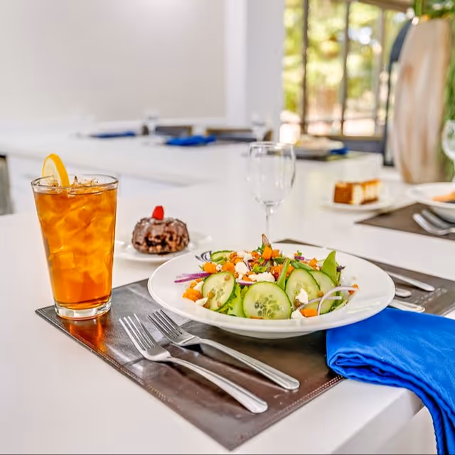 A plated cucumber salad, iced tea with a lemon slice, and a small dessert arranged on a set dining table.