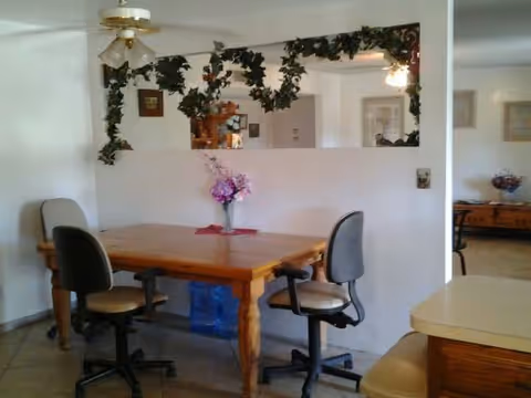 A dining area with a wooden table surrounded by four office-style chairs on wheels. Above the table, there is a ceiling fan with a light fixture. A large mirror on the wall is decorated with green leafy garlands. In the background, another room with a wooden sideboard and a vase of flowers is visible.