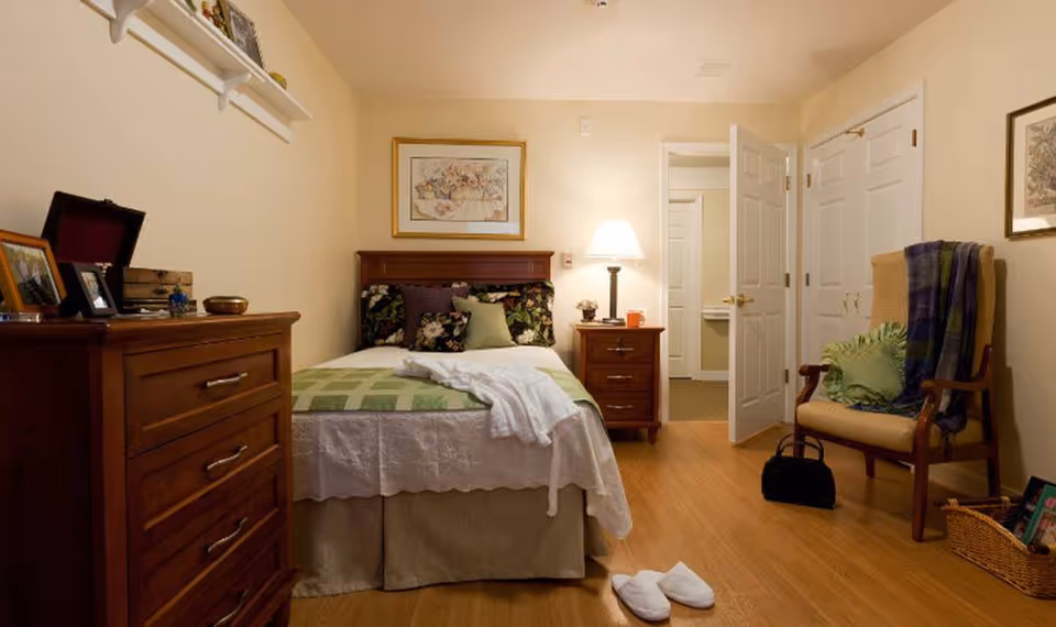 A cozy bedroom with a wooden bed featuring floral pillows and a green checkered blanket. There is a wooden dresser with framed photos and decorative items on the left side. A nightstand with a lamp and small decor is next to the bed. A cushioned chair with a green pillow and a plaid throw blanket is on the right side. The room has light-colored walls, wooden flooring, and an open door leading to another room. A pair of white slippers is placed on the floor near the bed.