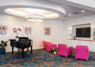 A bright and modern common area with a black grand piano, three pink armchairs, a flat-screen TV mounted on a white cabinet, colorful framed artwork on the wall, and a patterned carpet. There are also some potted plants adding greenery to the space.