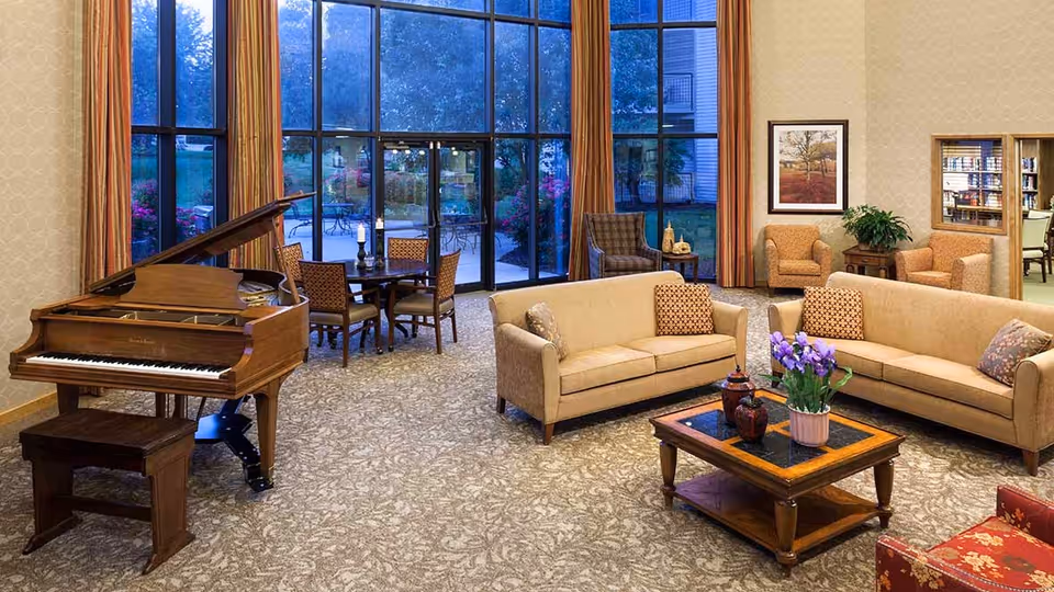 A spacious and well-lit living room area with large floor-to-ceiling windows showing an outdoor patio. The room features two beige sofas with patterned cushions, a wooden coffee table with a flower pot and decorative items, a wooden grand piano with a bench, a round table with four chairs, and additional armchairs near the walls. The walls are decorated with framed artwork and there are long curtains on the windows.