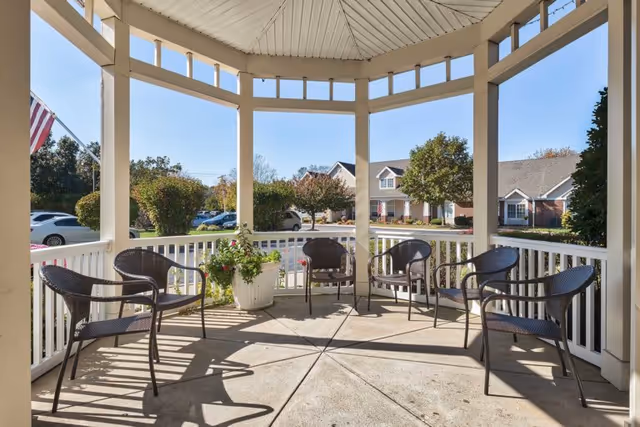 Outdoor covered seating area with six black wicker chairs arranged in a circle around a large white planter with flowers. The area is surrounded by white railings and overlooks a parking lot and residential-style buildings with trees and clear blue sky.