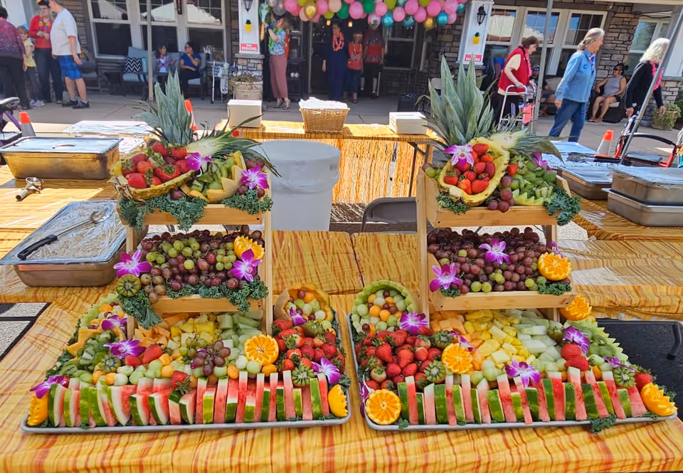 Two large trays of assorted fresh fruit arranged decoratively on a table with a yellow checkered tablecloth. The fruit includes watermelon slices, strawberries, grapes, pineapple, cantaloupe, honeydew melon, and orange slices, garnished with purple flowers and greenery. The trays are elevated on wooden stands. In the background, people are gathered outside a building decorated with colorful balloons.