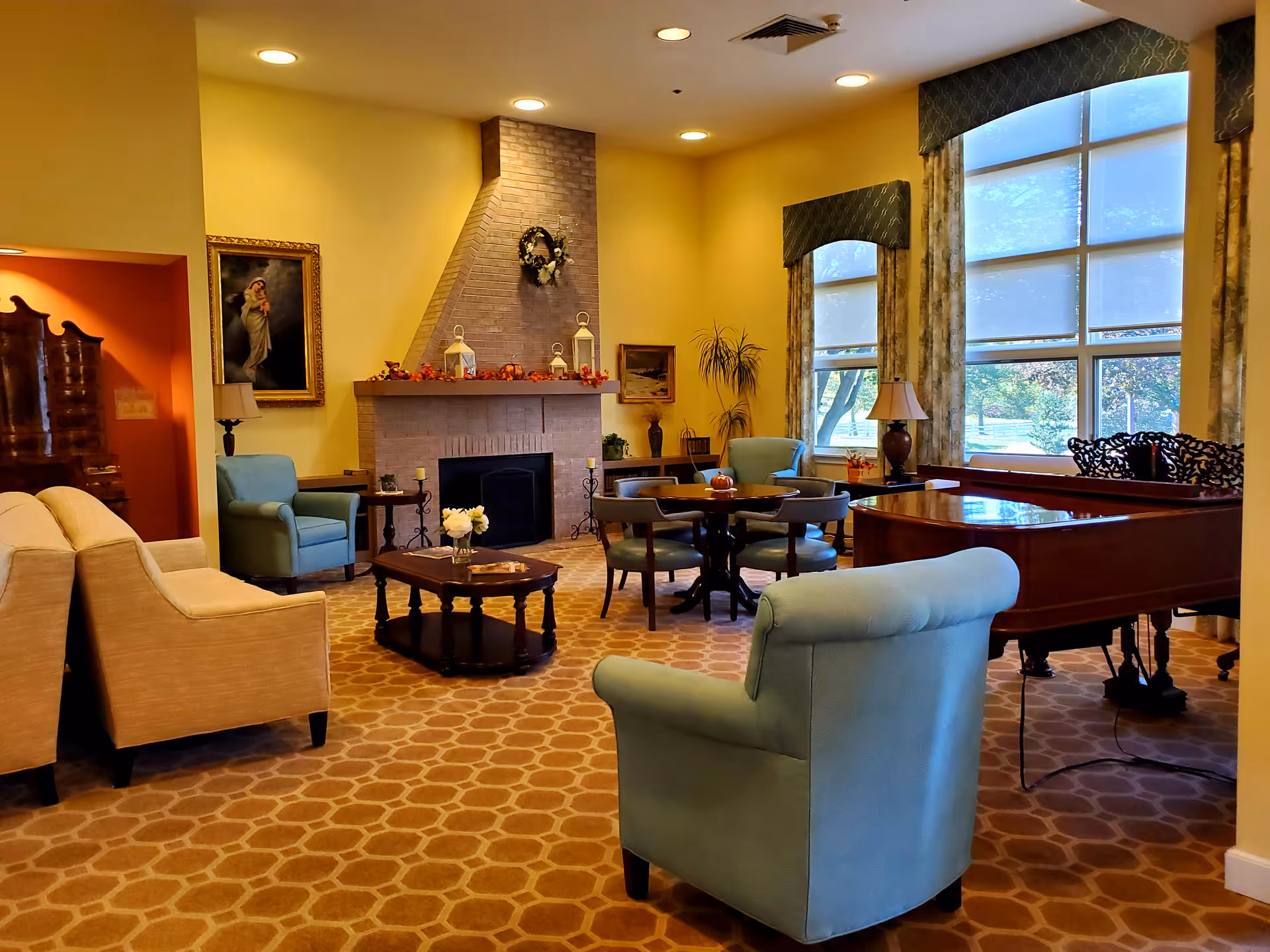 A warm communal living room with upholstered chairs, a fireplace, round tables, and a grand piano by large windows.