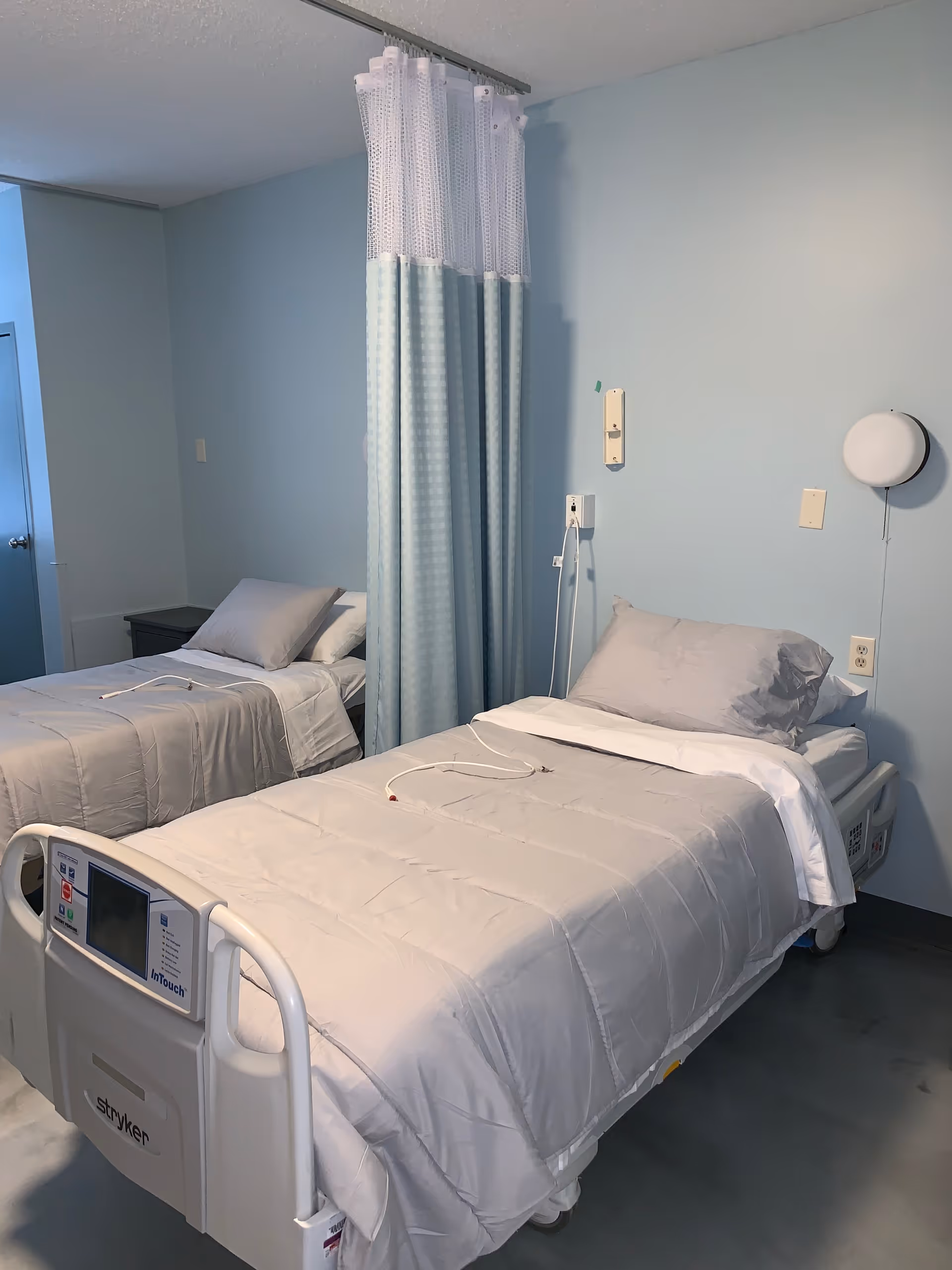 A hospital-style room with two single beds separated by a light blue privacy curtain. Each bed is made with light gray bedding and pillows. The bed in the foreground has a control panel labeled 'Stryker' and 'InTouch'. The walls are painted light blue, and there is a round wall light fixture above the bed on the right.