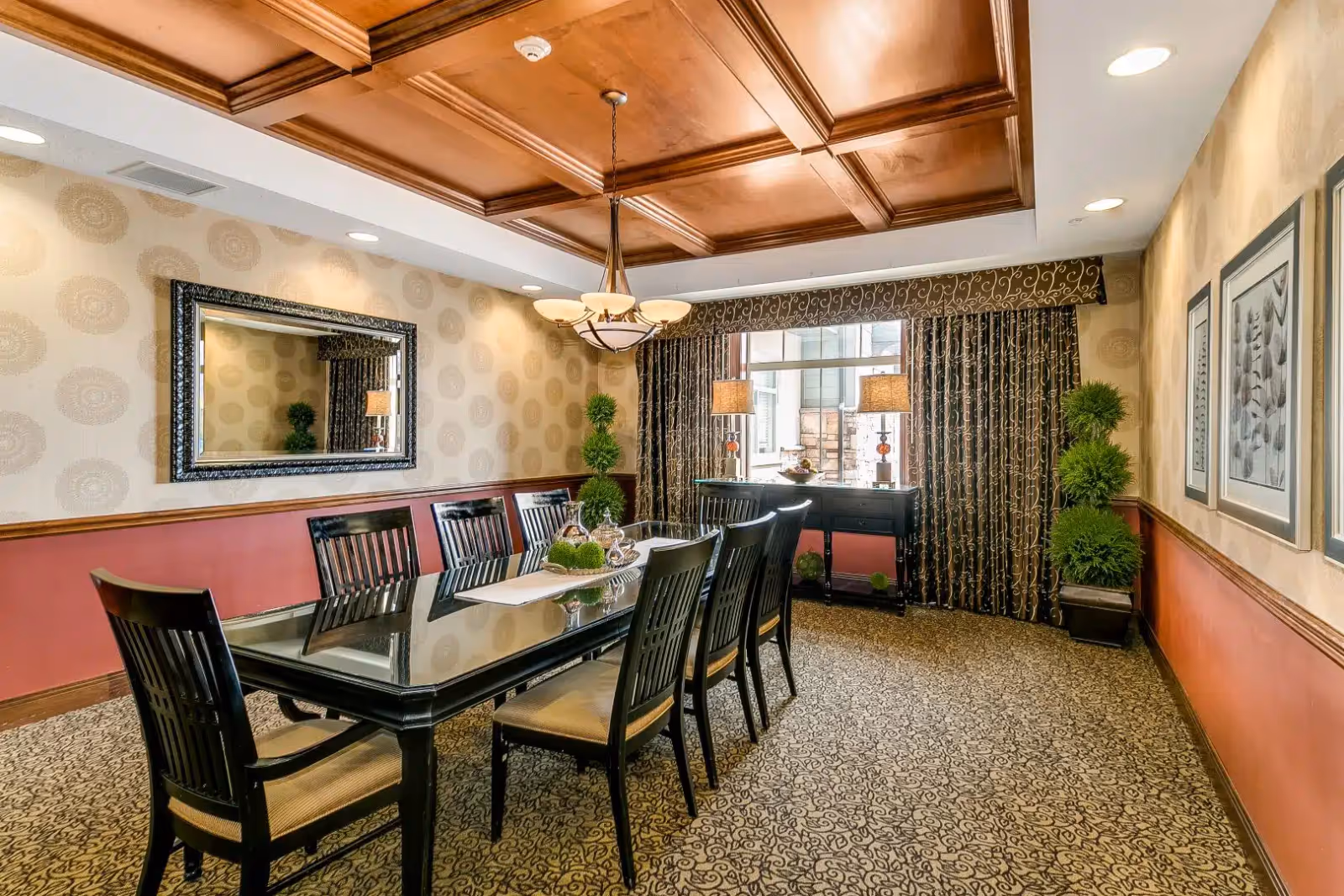 A formal dining room with a long rectangular glass-top table surrounded by eight black wooden chairs with beige cushions. The room features a patterned carpet, wallpaper with circular designs, a large decorative mirror on one wall, and three framed pictures on another. There is a wooden coffered ceiling with a chandelier hanging above the table. At the far end, a window with patterned curtains is flanked by two table lamps on a black sideboard, and two potted topiary plants are placed in the corners.