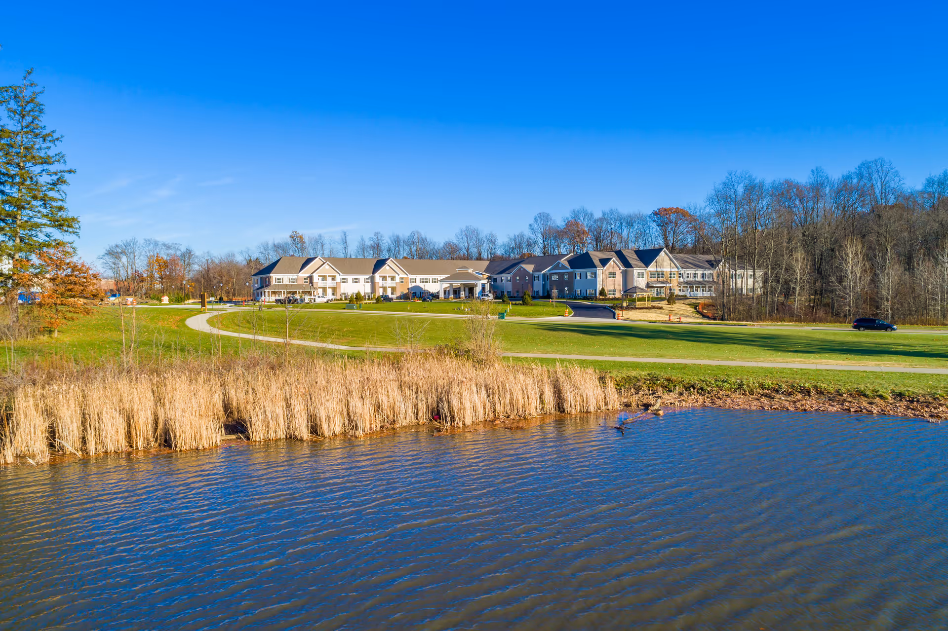 Wide exterior view of the Danbury Brunswick senior living facility with a large pond in the foreground, grassy area, and a clear blue sky. The building is surrounded by trees and a driveway with a parked car is visible.