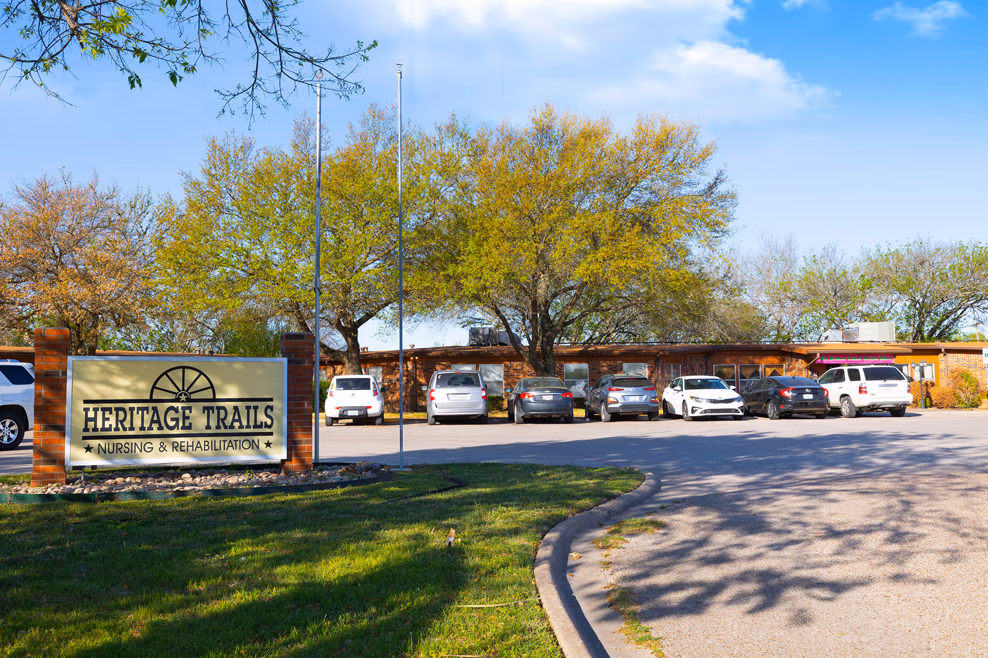 Exterior view of Heritage Trails Nursing & Rehabilitation facility with a sign in the foreground and several parked cars in front of a single-story brick building surrounded by trees under a blue sky.