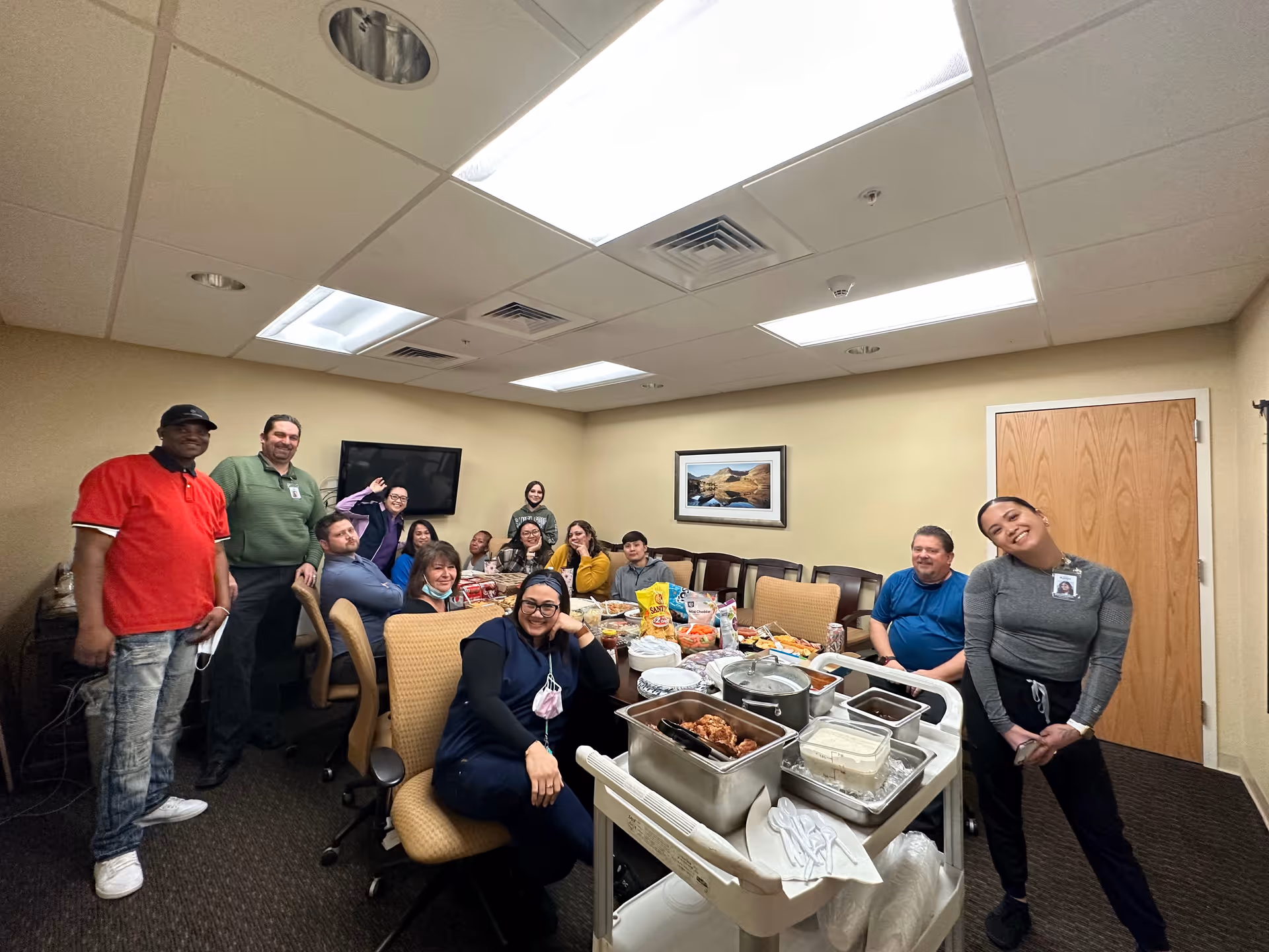 A group of people gathered in a conference room with beige walls and ceiling lights. They are seated around a table with various food items and snacks, smiling and posing for the photo. A TV is mounted on the wall, and a framed landscape picture hangs beside a wooden door.