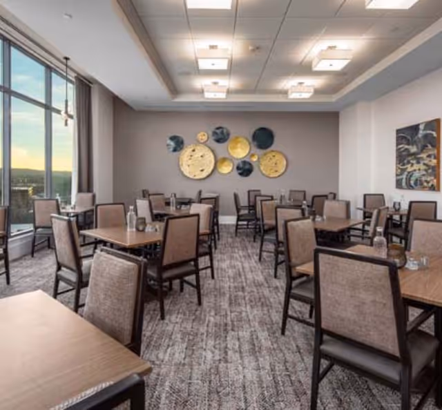 A dining room with multiple square wooden tables and cushioned chairs arranged neatly. The room has large windows on one side letting in natural light and offering a view outside. The walls are decorated with circular art pieces in gold and dark blue tones, and there is a carpeted floor with a patterned design.
