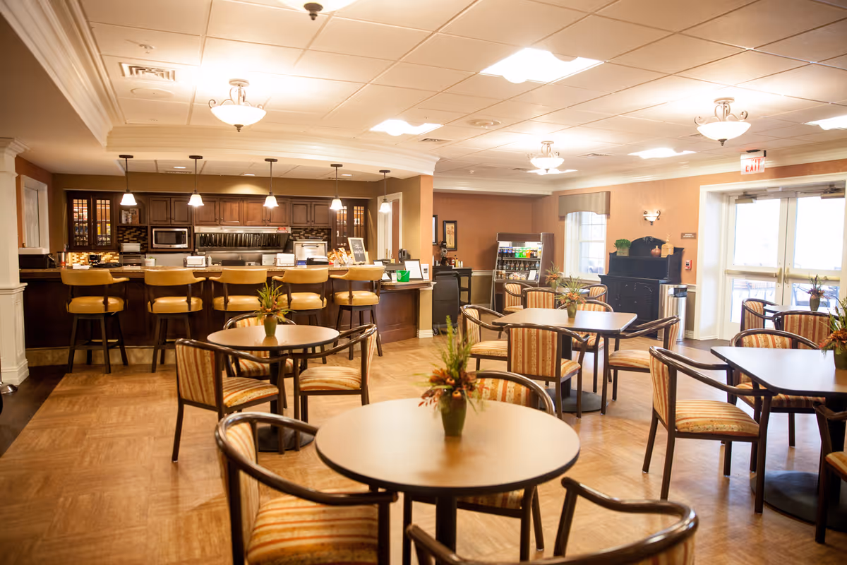 A warm and inviting dining area in a retirement community featuring several round tables with striped cushioned chairs. Each table has a small floral centerpiece. In the background, there is a kitchen counter with bar stools, pendant lights, and kitchen appliances including a microwave and toaster. The room has wood flooring, soft lighting, and large windows letting in natural light.