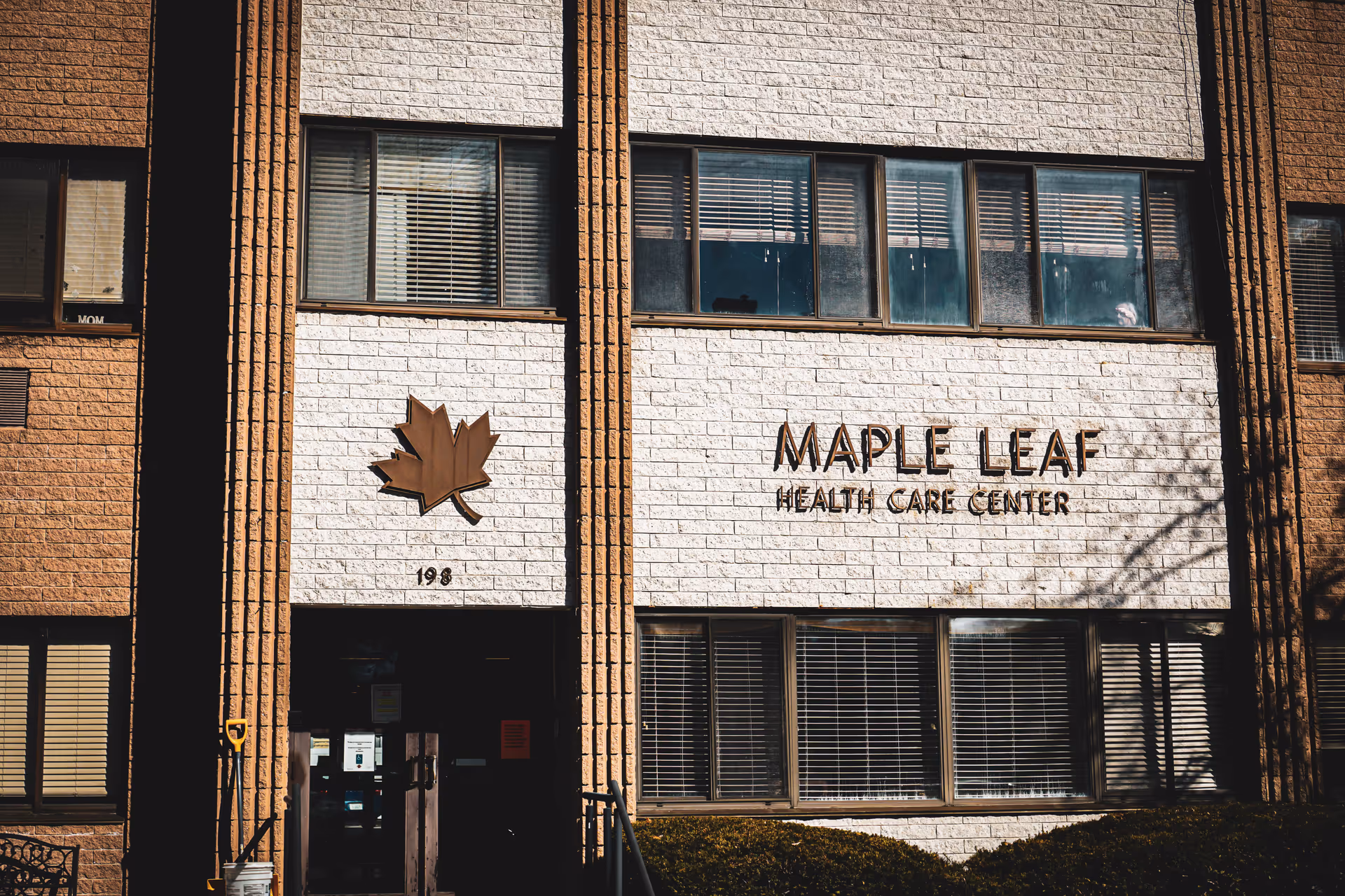 Exterior view of the Maple Leaf Health Care Center building showing a brick facade with windows and the facility's name and logo featuring a maple leaf mounted on the wall.