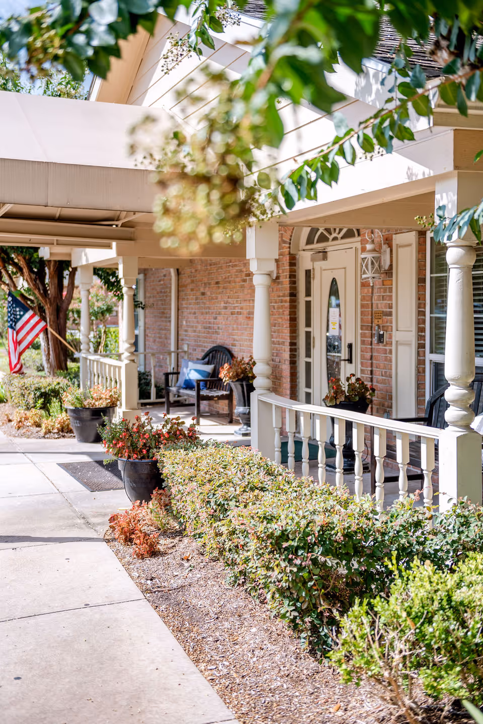 Front porch entrance with columns, potted plants, benches, and an American flag at a senior living facility.