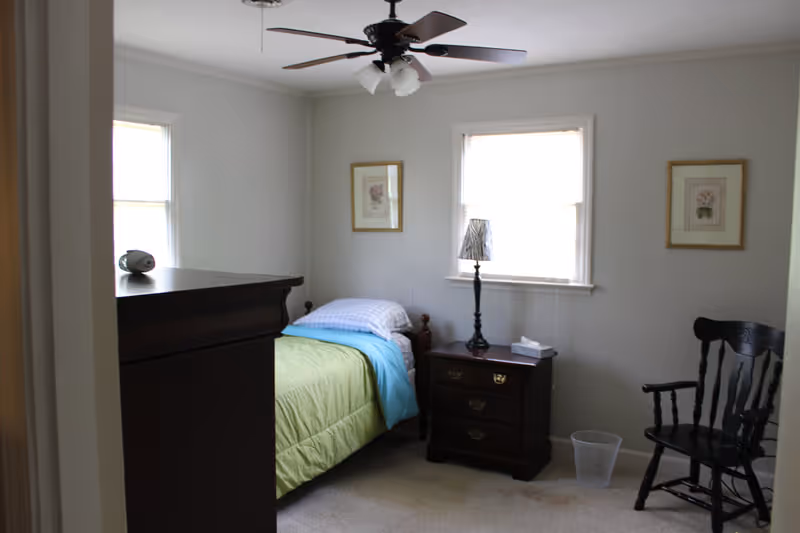 A small bedroom with a single bed covered with a green and blue blanket, a wooden nightstand with a lamp and tissue box, a wooden dresser, a black wooden chair, two framed floral pictures on the walls, and two windows letting in natural light.