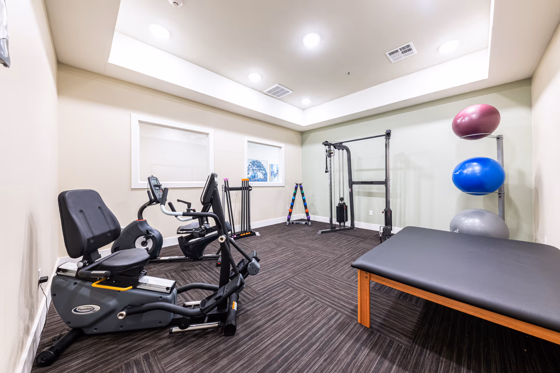 A small fitness room with exercise equipment including a recumbent bike, a multi-functional cable machine, a rack with colorful dumbbells, three large exercise balls stacked on a rack, and a padded exercise bench. The room has beige walls, a dark carpeted floor, and recessed ceiling lights.