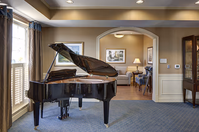 A spacious room with a black grand piano in the foreground, positioned on a blue carpet. Behind the piano, there is an arched doorway leading to a cozy sitting area with a sofa, armchairs, a side table with a lamp, and framed artwork on the walls. Large windows with curtains allow natural light into the room.