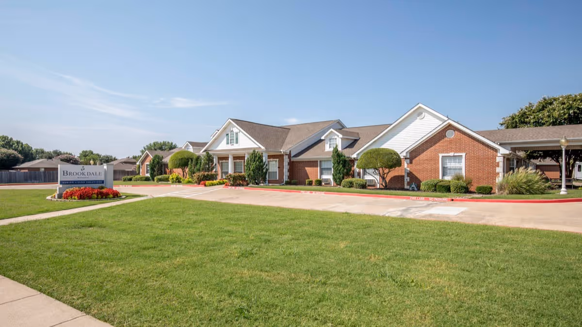 Exterior view of Brookdale Watauga senior living facility showing a single-story brick building with white trim, manicured bushes, and a well-maintained lawn under a clear blue sky. A sign with the facility name is visible near the driveway entrance.