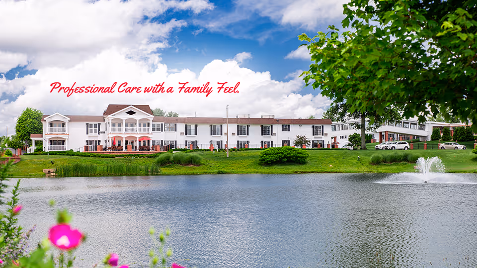 A large white nursing home building with multiple windows and balconies, situated behind a pond with a fountain. The sky is partly cloudy with blue patches, and there is greenery including trees and bushes around the pond. Text on the image reads 'Professional Care with a Family Feel.'